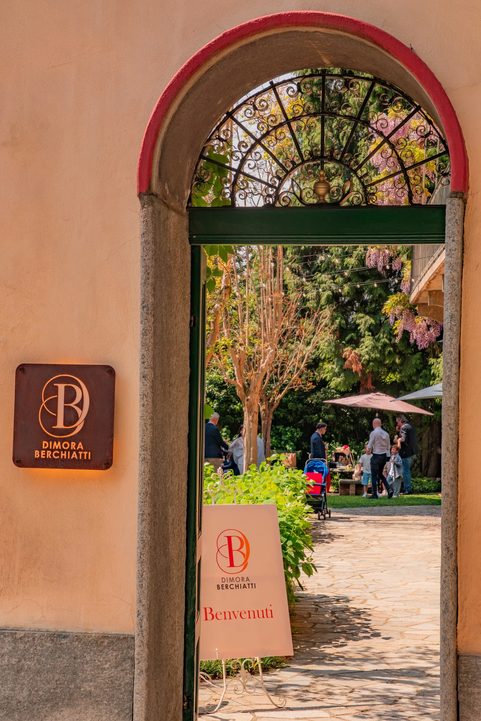L’ingresso dalla via: l’arco con gli stipiti di pietra e la lunetta in ferro battuto, la targa con il marchio e il giardino oltre la porta