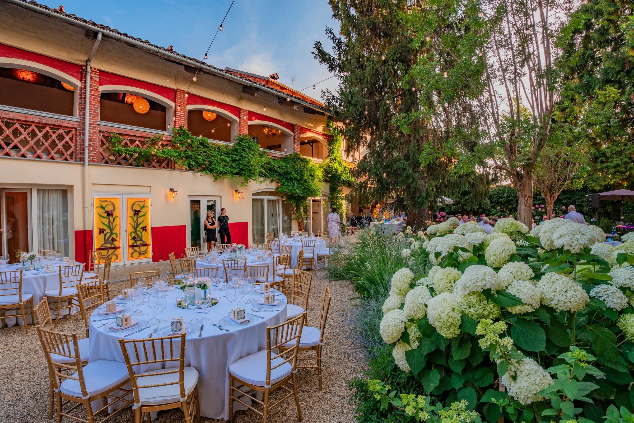 La corte apparecchiata per un matrimonio con i tavoli rotondi e le sedie dorate nell’ultima luce del giorno, fra le ortensie, con le arcate della sala superiore accese