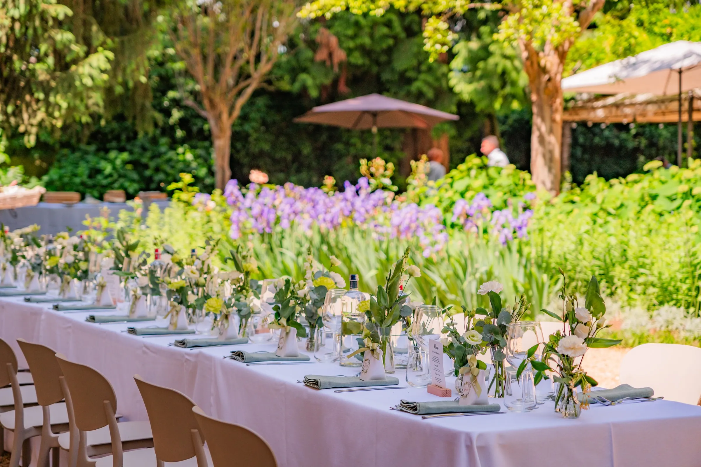 Il tavolo lungo del pranzo di una comunione apparecchiato in corte, con i vasetti di fiori bianchi e gli iris viola dell’aiuola dietro