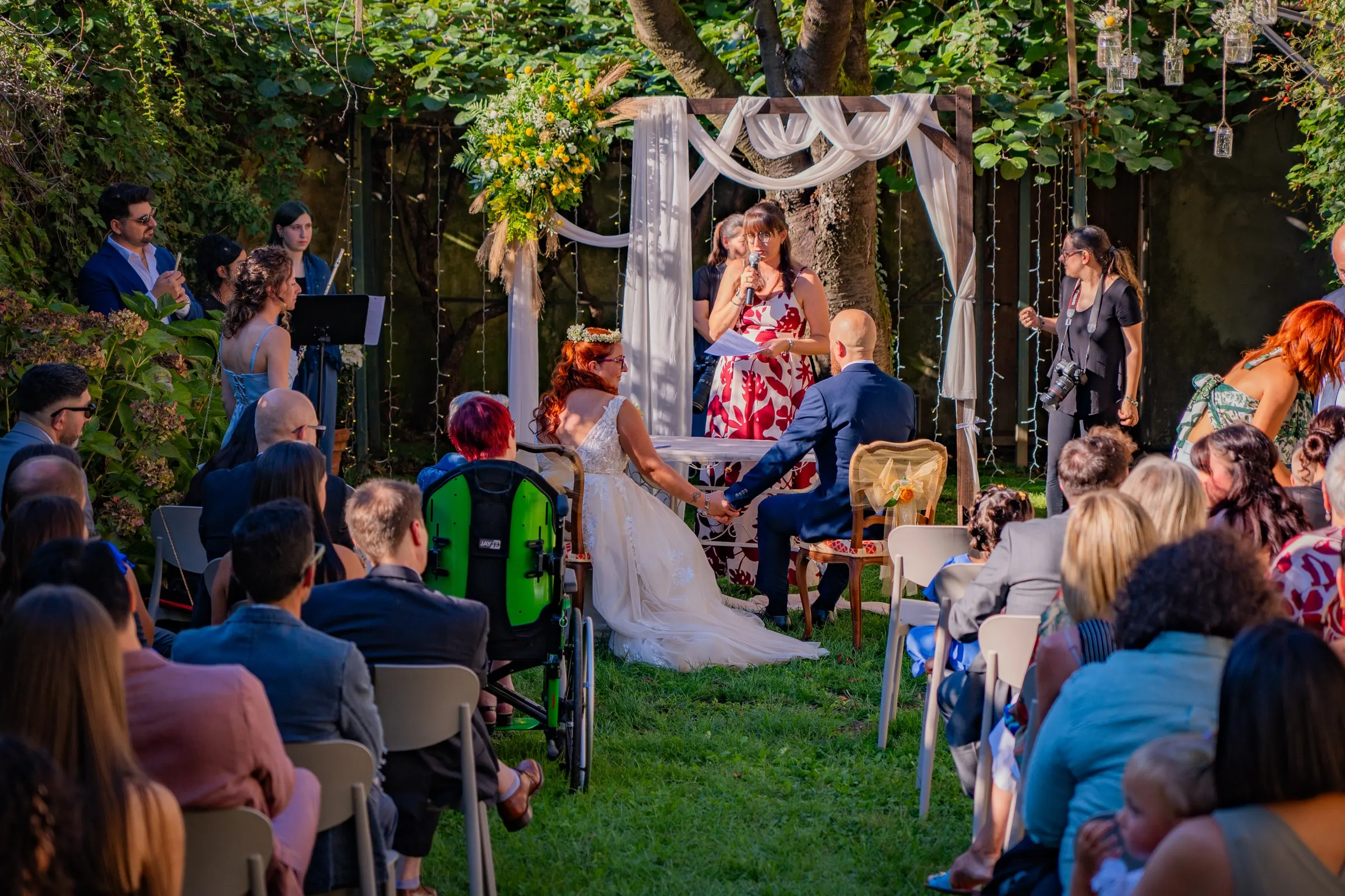 Il rito civile in giardino: gli sposi seduti sotto l’arco di tessuto bianco e fiori gialli, la celebrante in piedi davanti a loro e gli invitati sulle sedie sul prato