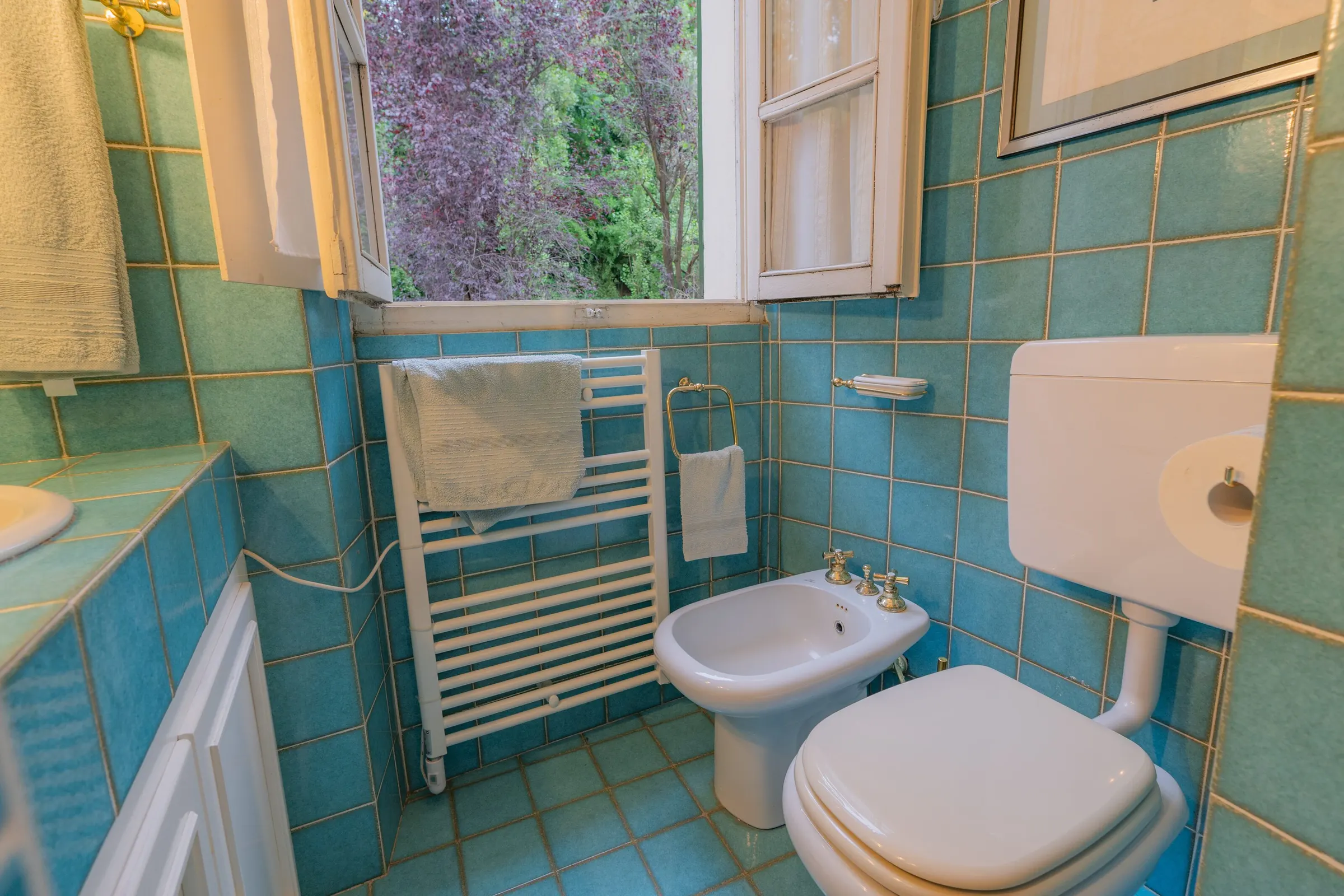La finestra del bagno turchese aperta sulle fronde del glicine, con il termoarredo e i sanitari