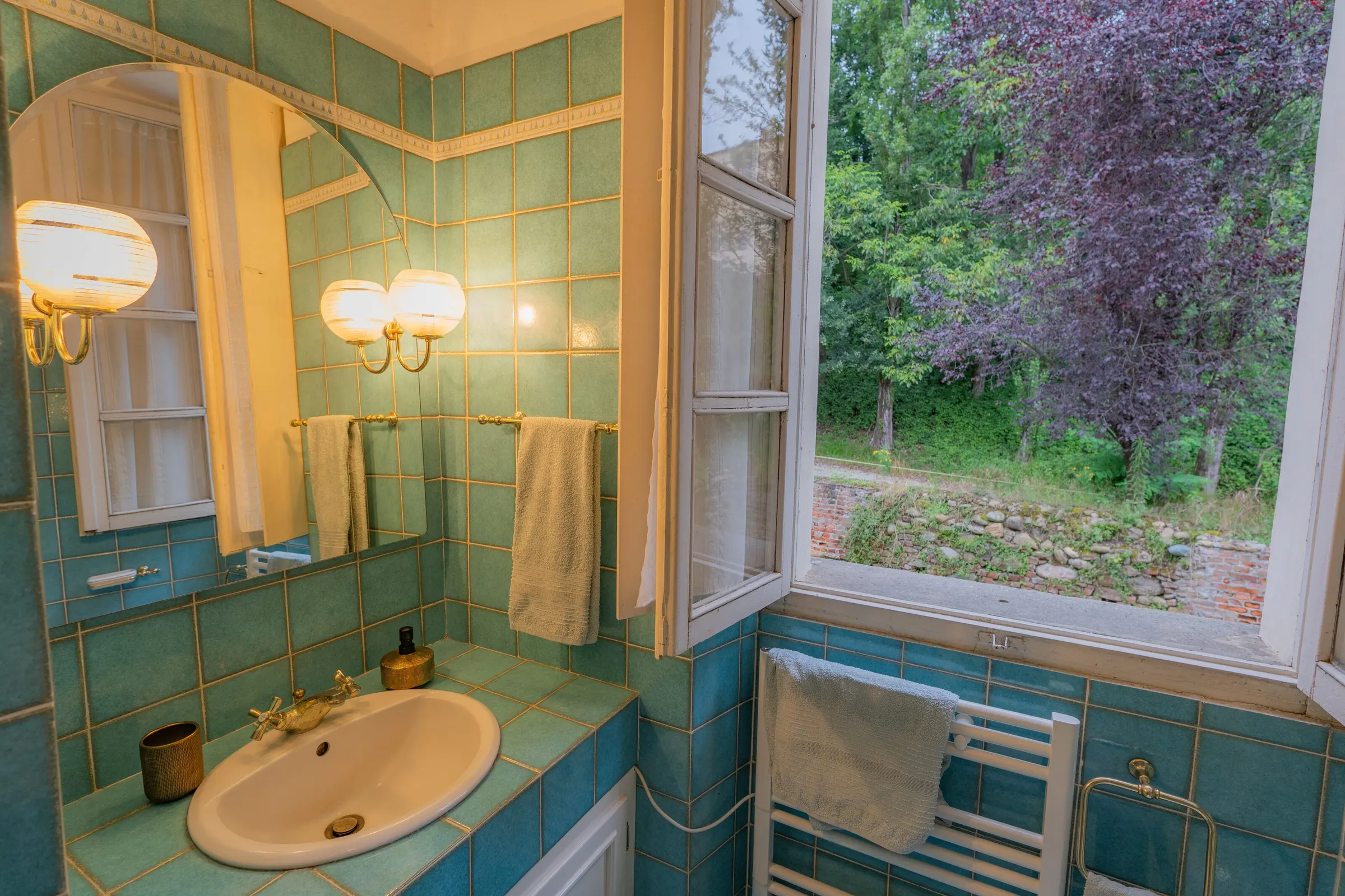 Il bagno rivestito di piastrelle turchesi, con il lavabo, lo specchio e la finestra spalancata sul verde
