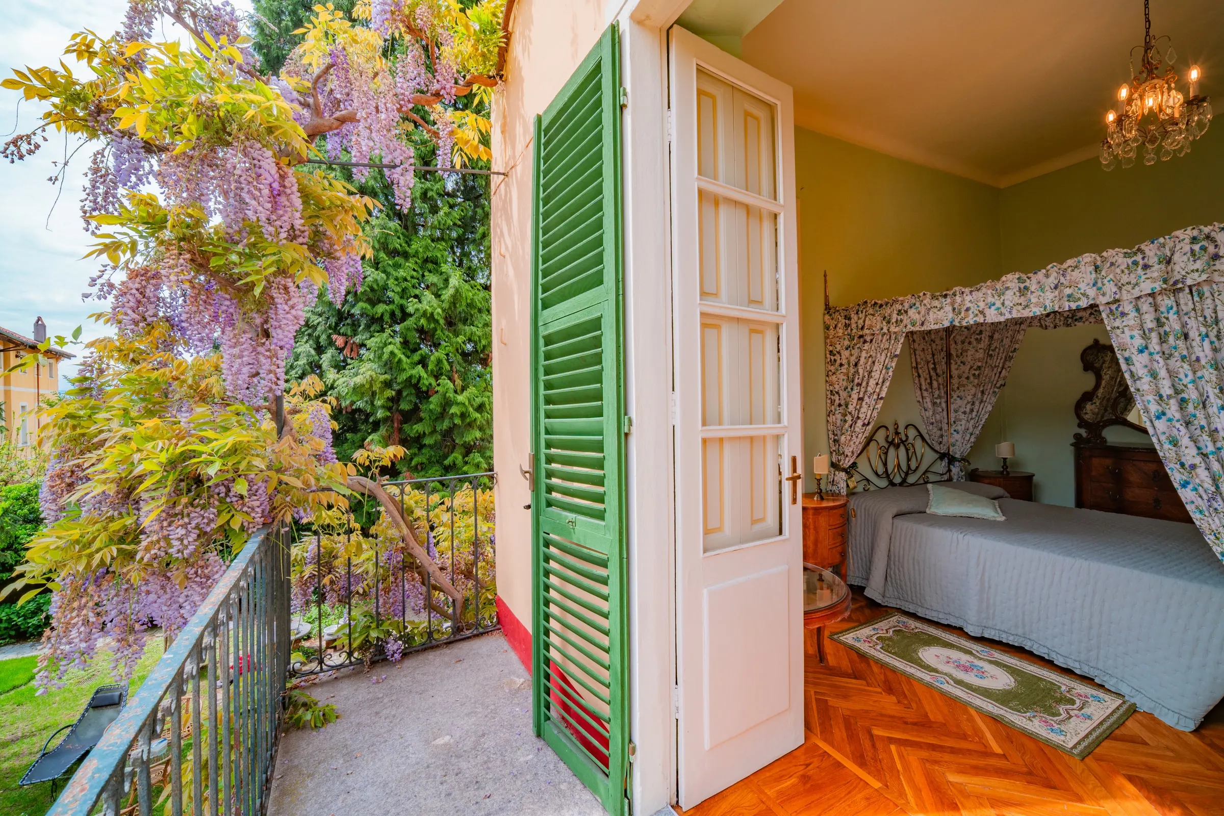 Il glicine in fiore visto dal balcone, con la persiana verde aperta e la camera del baldacchino oltre la portafinestra