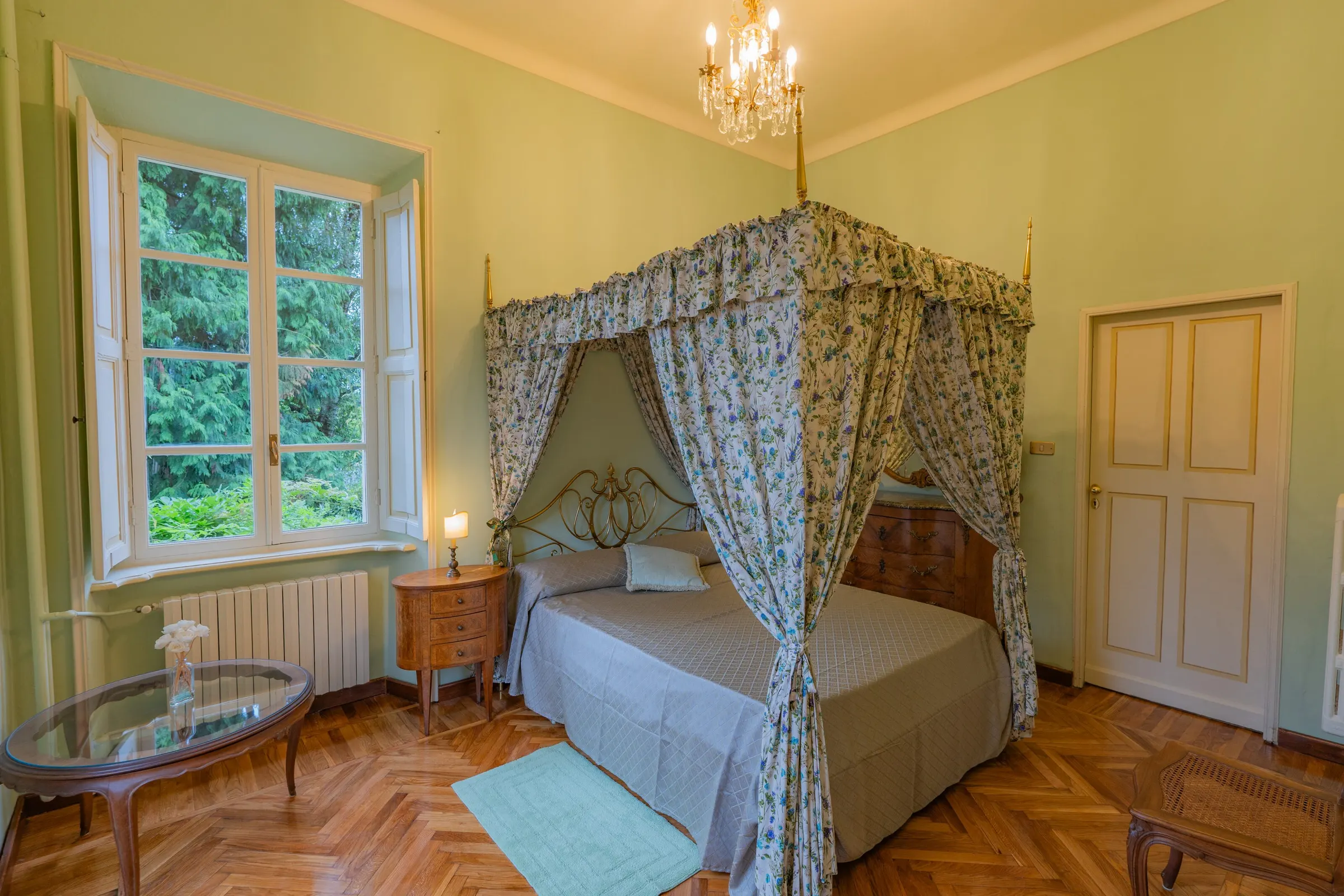 La camera col letto a baldacchino: le tende a fiori, la parete verde salvia, il parquet a spina di pesce e la finestra sul giardino