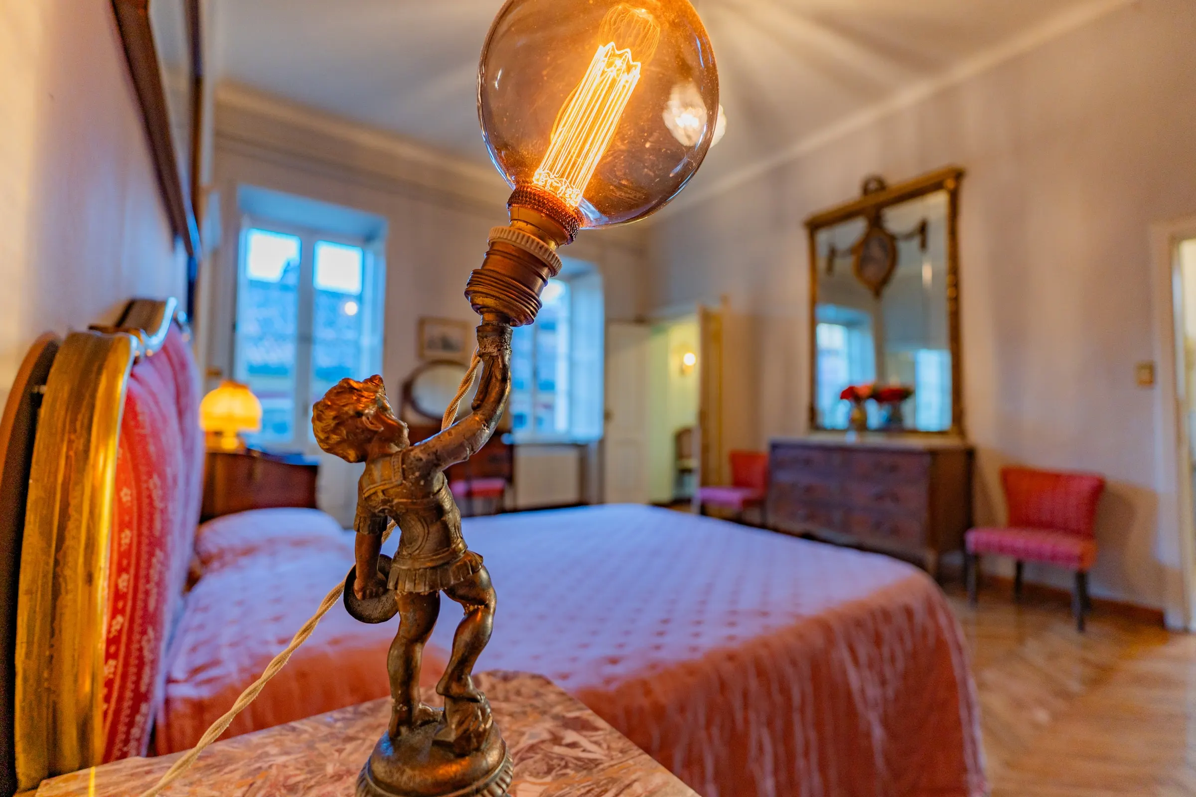 La lampada con il putto di metallo che regge la lampadina a filamento accesa; dietro, il letto e la specchiera della camera matrimoniale
