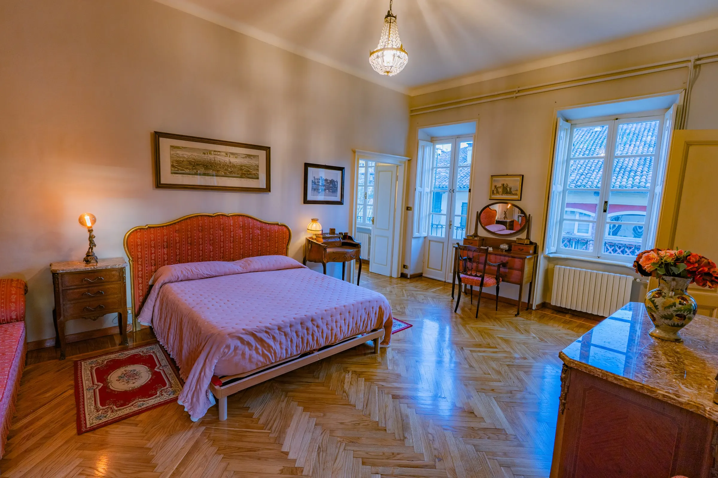 La camera matrimoniale con il letto dalla testata rosa, il parquet a spina di pesce, il lampadario di cristallo e le due finestre sui tetti