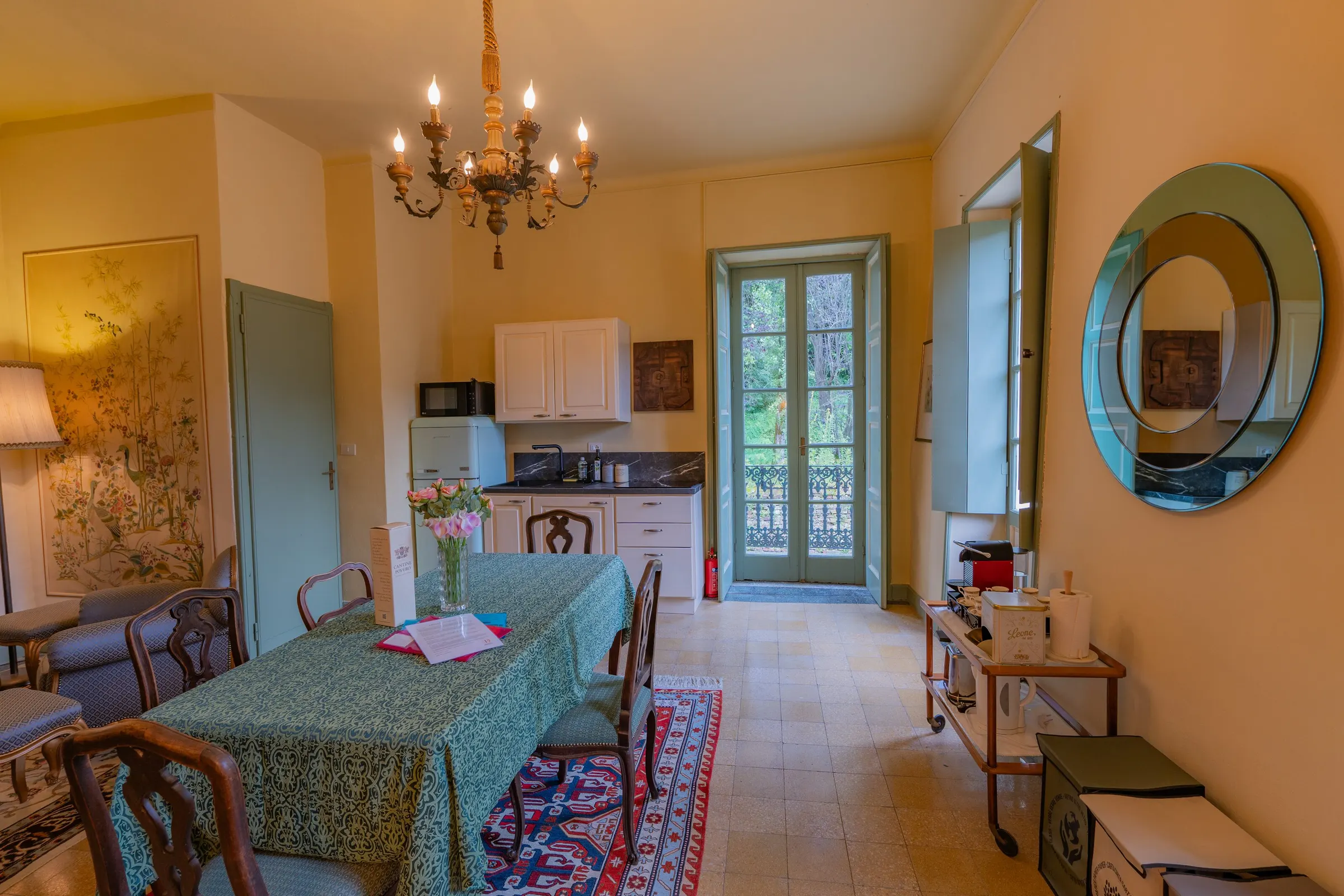 Il soggiorno con il tavolo da pranzo e la tovaglia verde, il tappeto persiano, il lampadario di ottone e la portafinestra sul balcone