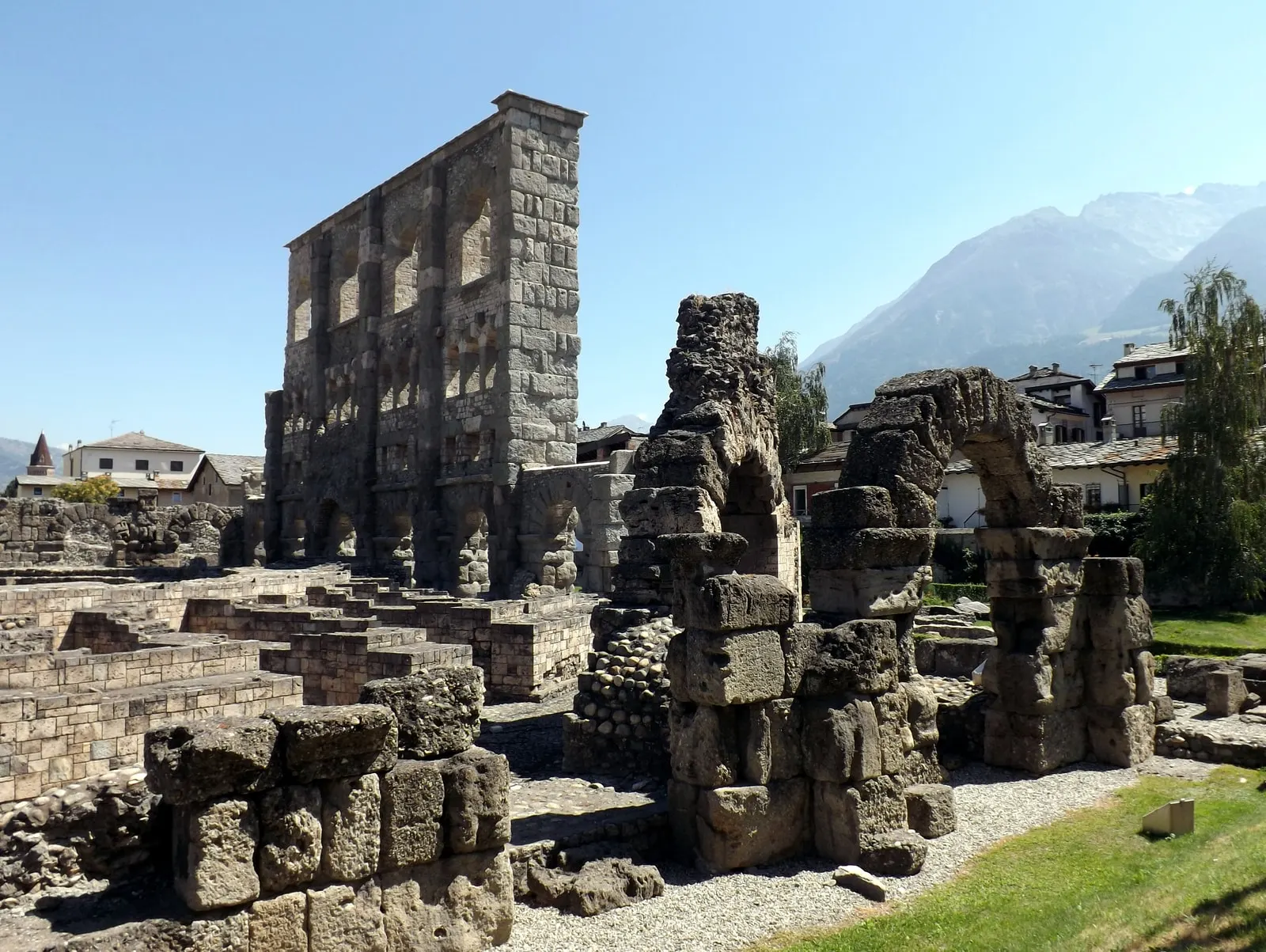 La facciata del teatro romano di Aosta, alta e aperta da archi, con i resti delle arcate in primo piano e le montagne sul fondo