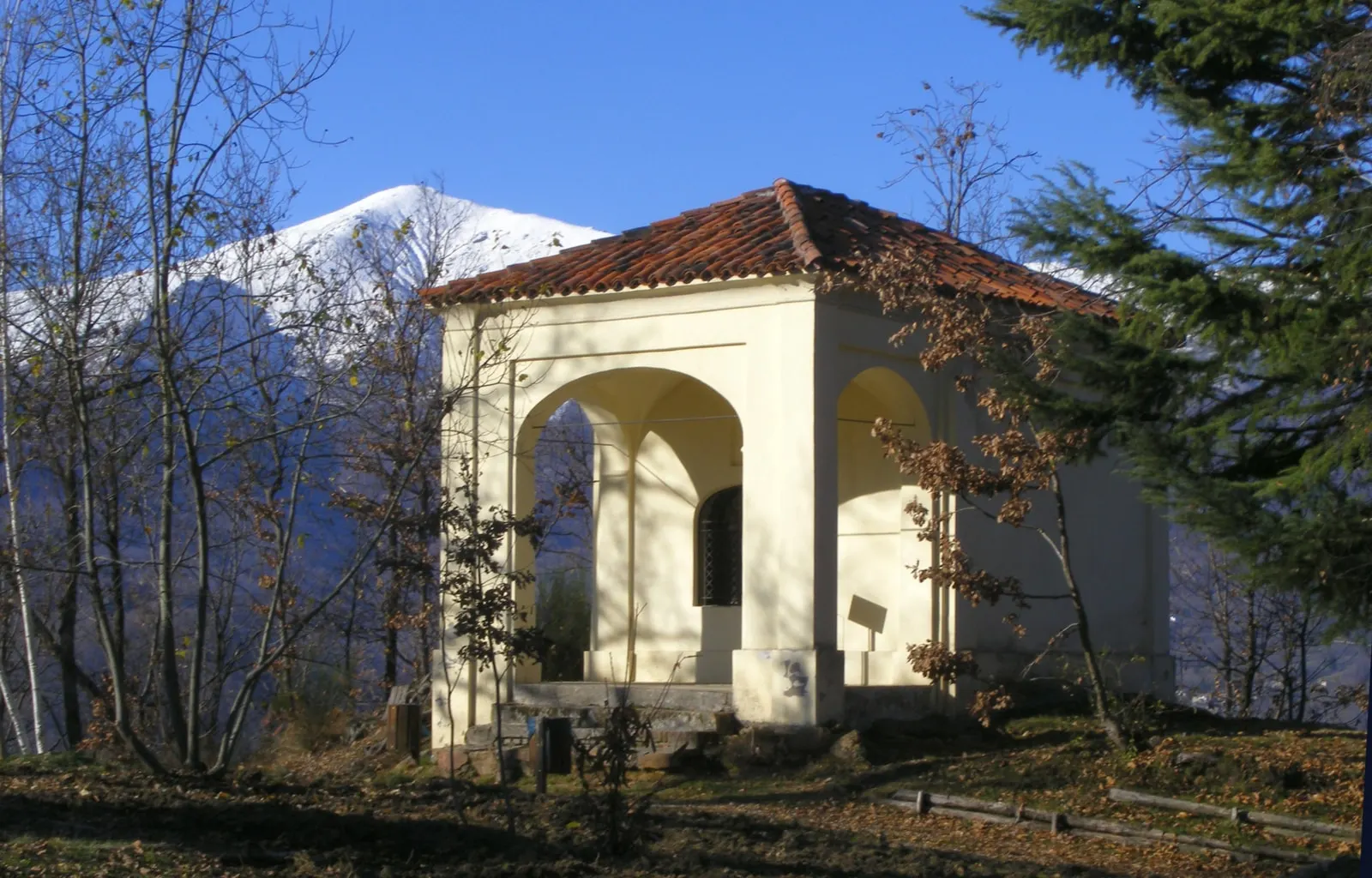 Una cappella del Sacro Monte di Belmonte, con gli archi e il tetto di coppi, fra gli alberi e una montagna innevata