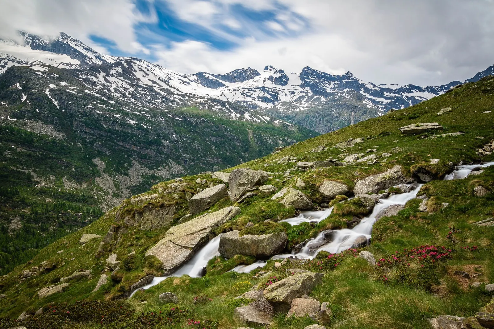 Un torrente che scende fra i massi e i prati della Valle dell’Orco, con le montagne innevate sullo sfondo